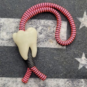 Peut inclure: Un cordon de cou sur le thème dentaire avec un charme de dent crème et un cordon tressé rose et noir. Le cordon est présenté sur un fond à motifs gris et blanc.