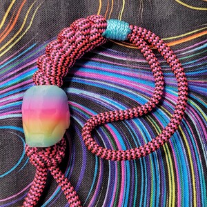 May include: A pink and black rope bracelet with a rainbow-coloured resin skull charm. The bracelet has gold-coloured metal beads at the ends.