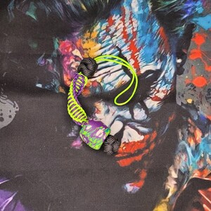 Puede incluir: Un llavero de paracord verde y morado con una cuenta negra y una cuenta verde y morada. El llavero está sobre un fondo negro y colorido.