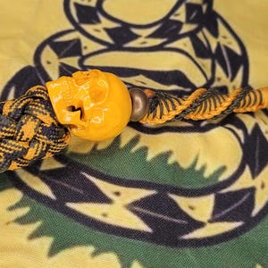 May include: A handmade lanyard with an orange skull bead, a braided black and yellow cord, and a black loop. The lanyard is crafted from durable materials and is designed for attaching to keys or other small items.