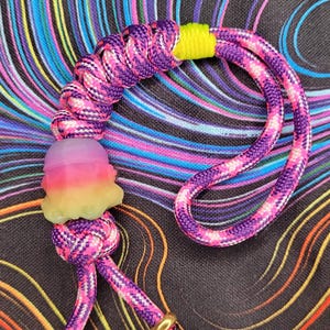 May include: A purple and pink braided rope bracelet with a yellow accent and a multi-colored resin bead. The bracelet is on a black and colorful background.