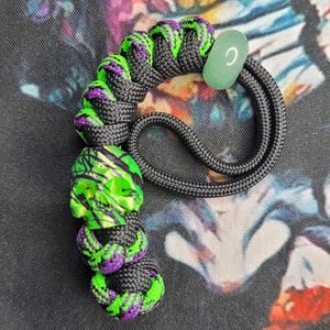 Peut inclure: Un cordon noir fait à la main avec une perle en forme de crâne vert et violet. Le cordon a des accents verts et violets. Une perle de pierre vert clair est attachée à l'extrémité.
