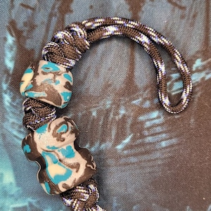 Op de afbeelding: Een zwart en blauw touw met twee blauwe en witte kralen. Het touw heeft een knoop aan de bovenkant en twee metalen ringen aan de onderkant.