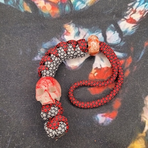 May include: A red, black, and grey paracord skull bead with a clear skull charm. The bead is accented with a small orange bead. The design is on a colourful patterned background.