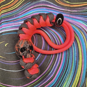 May include: A red and black paracord lanyard with a skull charm. The skull is black with red accents. The lanyard features a black and red woven design and a black, curved bead at the end. The background has a colorful, swirling pattern.