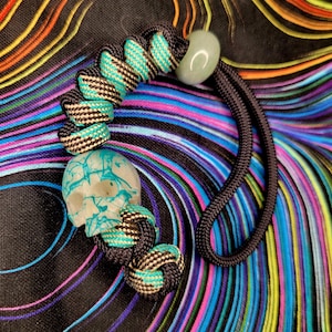 Peut inclure: Un cordon fait à la main avec une perle en forme de crâne ornée d'accents turquoise, reliée à une série de nœuds tissés noirs, turquoise et blancs. Une perle de couleur jade et un cordon noir complètent le design. Le fond présente un motif tourbillonnant coloré.