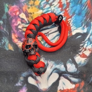 May include: A paracord lanyard featuring a black and red braided pattern with a detailed skull bead. The skull is black with red crackle-like markings. The cord is woven in a thick, textured knot design, ending in a loop with a small black bead.