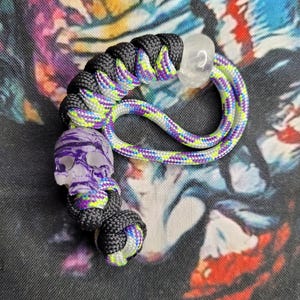May include: A handcrafted lanyard with a purple skull charm and a clear crystal bead. The lanyard is made of black and multicolored paracord in shades of purple, green, and yellow. The skull charm has a purple and white striped design.