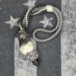 Op de afbeelding: Een zwart-wit paracord koord met een metalen bedel in de vorm van een pestmeestermasker. Het ontwerp bevat een gevlochten lus, een witte knoopkraal en een textuurrijke zwart-witte bolvormige kraal, verbonden aan het donkere metalen figuur.