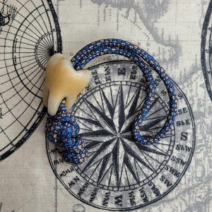 May include: A tooth-shaped pendant with a blue and tan cord. The pendant is a light tan colour. The cord is a dark blue with tan flecks. The pendant is lying on a map-themed fabric.