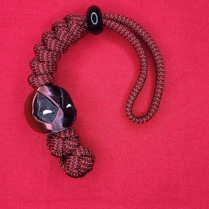 Puede incluir: Un cord&oacute;n rojo y negro con un dije de m&aacute;scara de Deadpool. El dije es negro con detalles de ojos blancos. El cord&oacute;n est&aacute; hecho de cord&oacute;n trenzado rojo y negro, con una cuenta y un lazo negros.