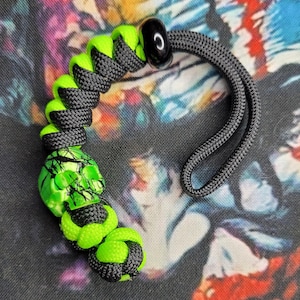 May include: A black and neon green paracord lanyard with a skull bead. The lanyard features a black cord with neon green accents and a skull-shaped bead. The lanyard has a black loop for attachment.