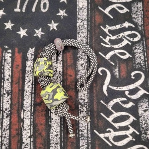 May include: A black and white paracord bracelet with a green and black camouflage bead and a silver bead. The bracelet is on a fabric background with the words "We the People" and "1776" printed on it.