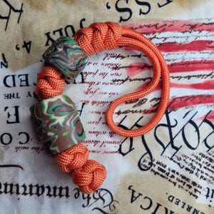 Op de afbeelding: Een oranje paracord lanyard met twee gemarmerde kralen in tinten bruin, groen en wit. De lanyard heeft een lus en decoratieve knopen. De achtergrond is een stof met patriottische tekst en afbeeldingen.