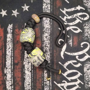May include: A black paracord bracelet featuring two square beads with a marbled pattern of gray and neon yellow. The bracelet includes a small, round, tan bead and gold-colored metal accents.