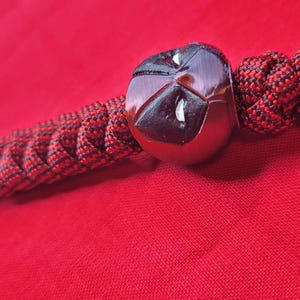 Puede incluir: Un llavero hecho a mano con una cuenta esf&eacute;rica oscura y reflectante y un cord&oacute;n trenzado rojo y negro. El llavero tiene un lazo para sujetar llaves y un nudo decorativo. El fondo es de color rojo liso.