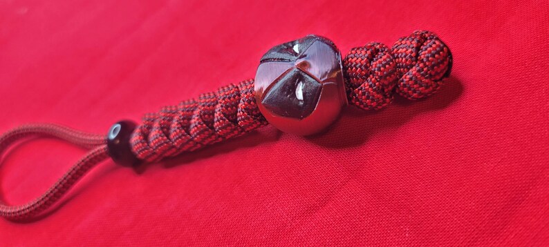 Puede incluir: Un llavero hecho a mano con una cuenta esf&eacute;rica oscura y reflectante y un cord&oacute;n trenzado rojo y negro. El llavero tiene un lazo para sujetar llaves y un nudo decorativo. El fondo es de color rojo liso.