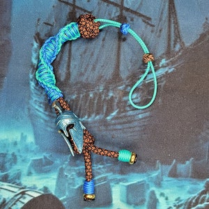 Op de afbeelding: Een blauwe en groene paracord sleutelhanger met een bruine knoop en een zilveren helmbedel. De sleutelhanger heeft blauwe en groene kralen.