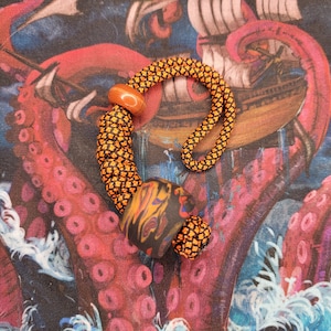 May include: A black and orange lanyard with a wooden bead and a black and orange swirl bead. The lanyard is on a colourful fabric background.