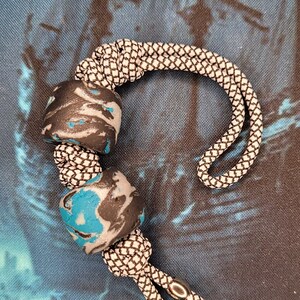 May include: A handmade lanyard featuring two marbled beads in black, grey, and turquoise, strung on a black and white braided cord. The cord is looped and finished with silver-toned metal accents.