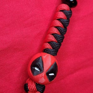 K&ouml;nnte beinhalten: Ein rotes und schwarzes Schl&uuml;sselband mit einem Deadpool-Design. Das Schl&uuml;sselband hat eine schwarze Kordel und eine Reihe von rot-schwarzen gewebten Abschnitten. Der Deadpool-Kopf ist rot mit schwarzen Akzenten und wei&szlig;en Augen.