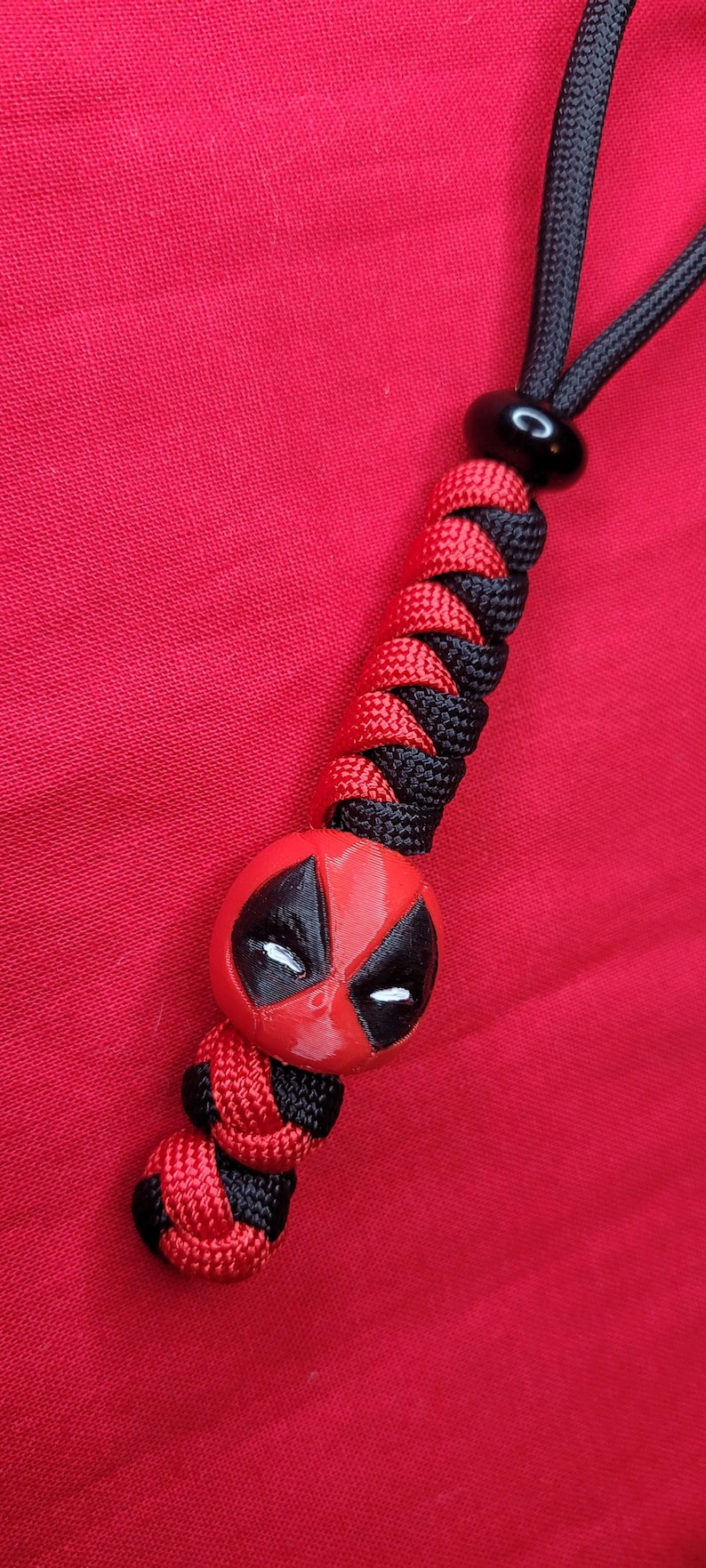 K&ouml;nnte beinhalten: Ein rotes und schwarzes Schl&uuml;sselband mit einem Deadpool-Design. Das Schl&uuml;sselband hat eine schwarze Kordel und eine Reihe von rot-schwarzen gewebten Abschnitten. Der Deadpool-Kopf ist rot mit schwarzen Akzenten und wei&szlig;en Augen.