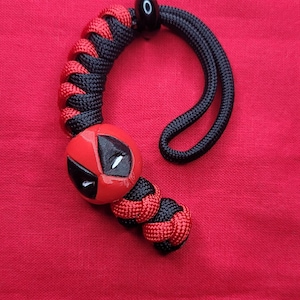 K&ouml;nnte beinhalten: Ein roter und schwarzer Paracord-Schl&uuml;sselanh&auml;nger mit Deadpool-Design. Der Schl&uuml;sselanh&auml;nger hat ein rot-schwarzes geflochtenes Muster, eine schwarze Schlaufe und einen roten Deadpool-Kopf mit wei&szlig;en Augen.