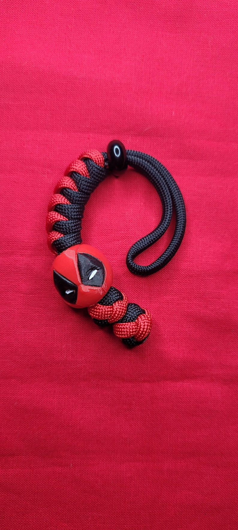 K&ouml;nnte beinhalten: Ein roter und schwarzer Paracord-Schl&uuml;sselanh&auml;nger mit Deadpool-Design. Der Schl&uuml;sselanh&auml;nger hat ein rot-schwarzes geflochtenes Muster, eine schwarze Schlaufe und einen roten Deadpool-Kopf mit wei&szlig;en Augen.