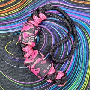 May include: A black and pink paracord bracelet with two black and pink marbled beads. The bracelet is on a black and rainbow swirl background.