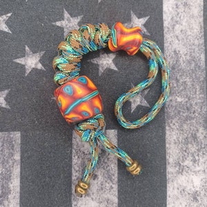 May include: A braided paracord bracelet with two orange, red, and blue glass beads. The bracelet is tied with a knot and has two gold metal beads at the end.