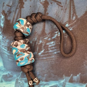 May include: A black lanyard with a decorative bead set. The beads are cylindrical and feature a pattern of blue, white, and black swirls. The lanyard is made of a woven black cord and has silver-colored metal accents.