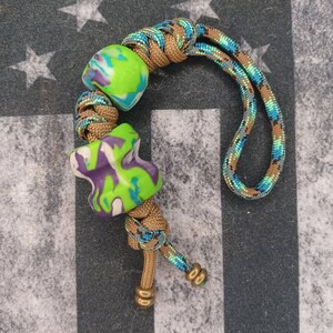 Op de afbeelding: Een groene, paarse en witte gepatenteerde kraal met een bruin en blauw paracord eromheen gewikkeld. Het paracord heeft gouden metalen kralen aan het einde.