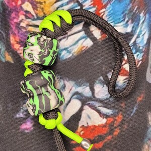May include: A black and green paracord bracelet with two green and black patterned beads. The bracelet has a green cord and a black cord with a knot at the end.