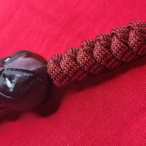 Puede incluir: Un cord&oacute;n de paracord negro y rojo con una cuenta negra y un elemento decorativo negro. El elemento decorativo tiene un dise&ntilde;o de cara. El cord&oacute;n est&aacute; sobre una superficie roja.