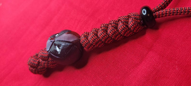 Puede incluir: Un cord&oacute;n de paracord negro y rojo con una cuenta negra y un elemento decorativo negro. El elemento decorativo tiene un dise&ntilde;o de cara. El cord&oacute;n est&aacute; sobre una superficie roja.