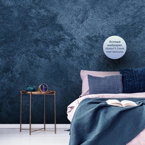 May include: A teal blue bed with a white blanket and a book on top. The bed has a pink headboard and pillows. A gold side table with a green decorative object and a clock is next to the bed. The wall is a dark blue textured wallpaper. The text on the wall reads "Printed wallpaper, doesn't have real texture."
