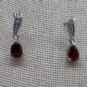 May include: A pair of silver-toned drop earrings. Each earring features a teardrop-shaped, deep red gemstone suspended from a curved, embellished top with small, clear stones. The earrings are displayed on a textured, neutral-toned surface.