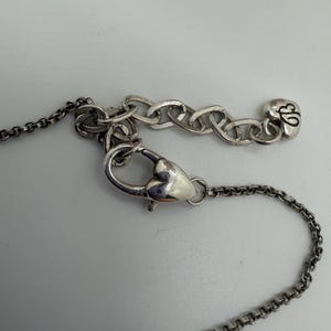 May include: Close-up of a silver necklace featuring a heart-shaped clasp and a chain. The necklace includes a small charm with a stylized design. The background is a plain, light grey surface.