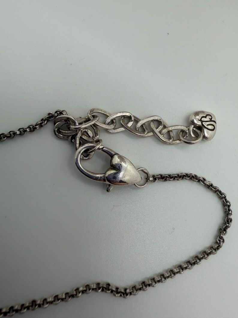 May include: Close-up of a silver necklace featuring a heart-shaped clasp and a chain. The necklace includes a small charm with a stylized design. The background is a plain, light grey surface.