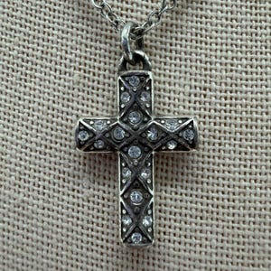 Puede incluir: Un collar con colgante de cruz plateada. La cruz est&aacute; adornada con numerosas piedras preciosas redondas y transparentes engastadas en un patr&oacute;n detallado. La cadena es de dise&ntilde;o delicado y eslabonado. El fondo es una tela texturizada de tono neutro.