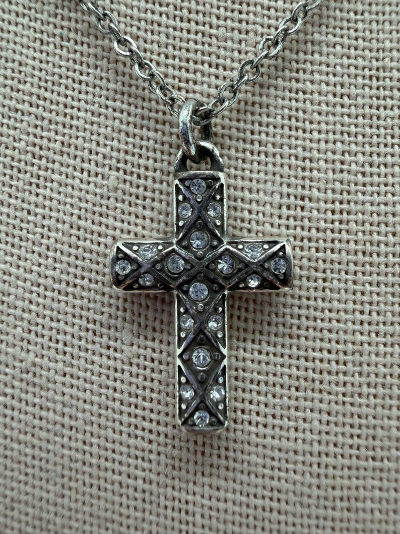 Puede incluir: Un collar con colgante de cruz plateada. La cruz est&aacute; adornada con numerosas piedras preciosas redondas y transparentes engastadas en un patr&oacute;n detallado. La cadena es de dise&ntilde;o delicado y eslabonado. El fondo es una tela texturizada de tono neutro.