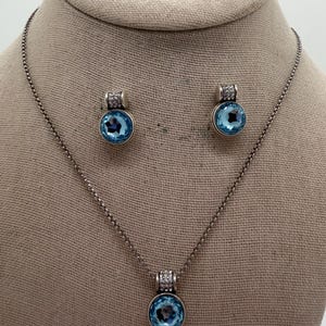 May include: A silver-coloured jewellery set comprising a necklace and matching earrings. Each piece features a round, light blue gemstone pendant with a halo of small, clear stones. The necklace and earrings are displayed on a neutral-toned surface.