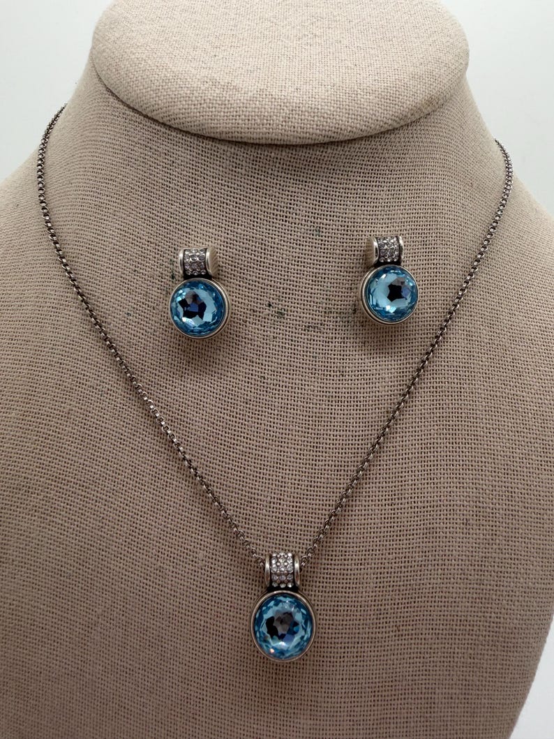 May include: A silver-coloured jewellery set comprising a necklace and matching earrings. Each piece features a round, light blue gemstone pendant with a halo of small, clear stones. The necklace and earrings are displayed on a neutral-toned surface.