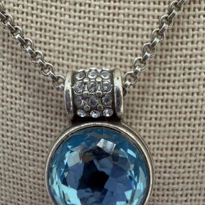 May include: Silver necklace featuring a round, light blue, faceted pendant. The pendant is set in a silver bezel and is attached to a silver chain. The top of the pendant has a silver band with small, clear crystals.