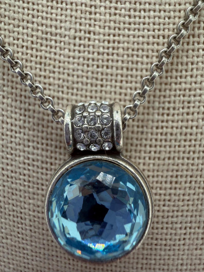 May include: Silver necklace featuring a round, light blue, faceted pendant. The pendant is set in a silver bezel and is attached to a silver chain. The top of the pendant has a silver band with small, clear crystals.