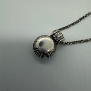 May include: A silver pendant necklace. The pendant is a small, round charm with horizontal grooves and a crystal-studded bail. The pendant hangs from a delicate chain. The jewellery is suitable for everyday wear. The necklace is a classic and elegant design.