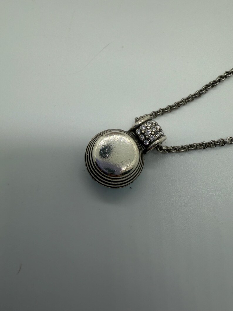 May include: A silver pendant necklace. The pendant is a small, round charm with horizontal grooves and a crystal-studded bail. The pendant hangs from a delicate chain. The jewellery is suitable for everyday wear. The necklace is a classic and elegant design.