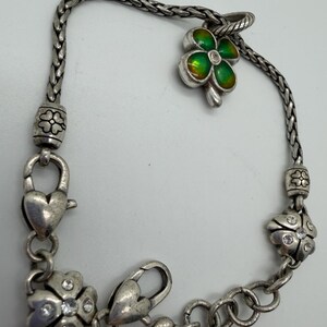 May include: Silver-toned bracelet featuring a four-leaf clover charm with green and yellow enamel, accented with a small clear crystal. The bracelet includes heart-shaped clasps and flower-shaped charms with crystal accents.