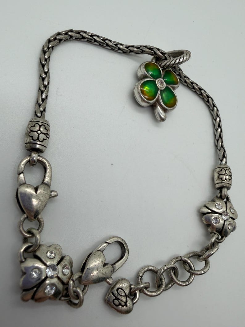 May include: Silver-toned bracelet featuring a four-leaf clover charm with green and yellow enamel, accented with a small clear crystal. The bracelet includes heart-shaped clasps and flower-shaped charms with crystal accents.