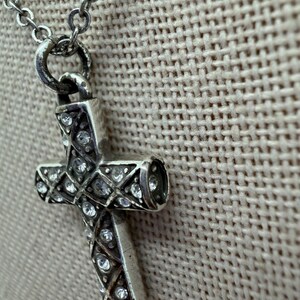 Puede incluir: Collar con colgante de cruz plateada y cadena fina. La cruz est&aacute; adornada con cristales transparentes dispuestos en forma de diamante. El collar se muestra sobre un fondo neutro y texturizado.